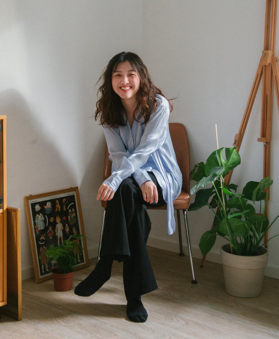 Sunny Chen sitting on a chair in a bright room with plants, an easel, and a framed vintage print